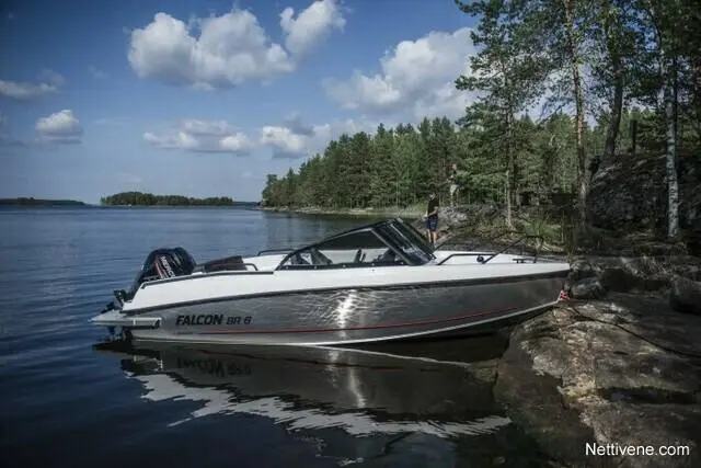 A Falcon SR 6 boat is moored by a rocky shore, with a person standing by the water amidst lush green trees and clouds.